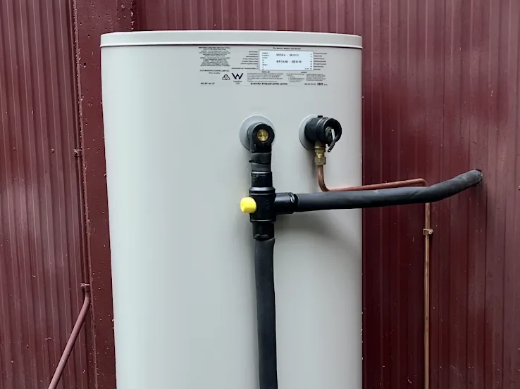A newly installed electric hot water cylinder with insulated pipework against a red fence