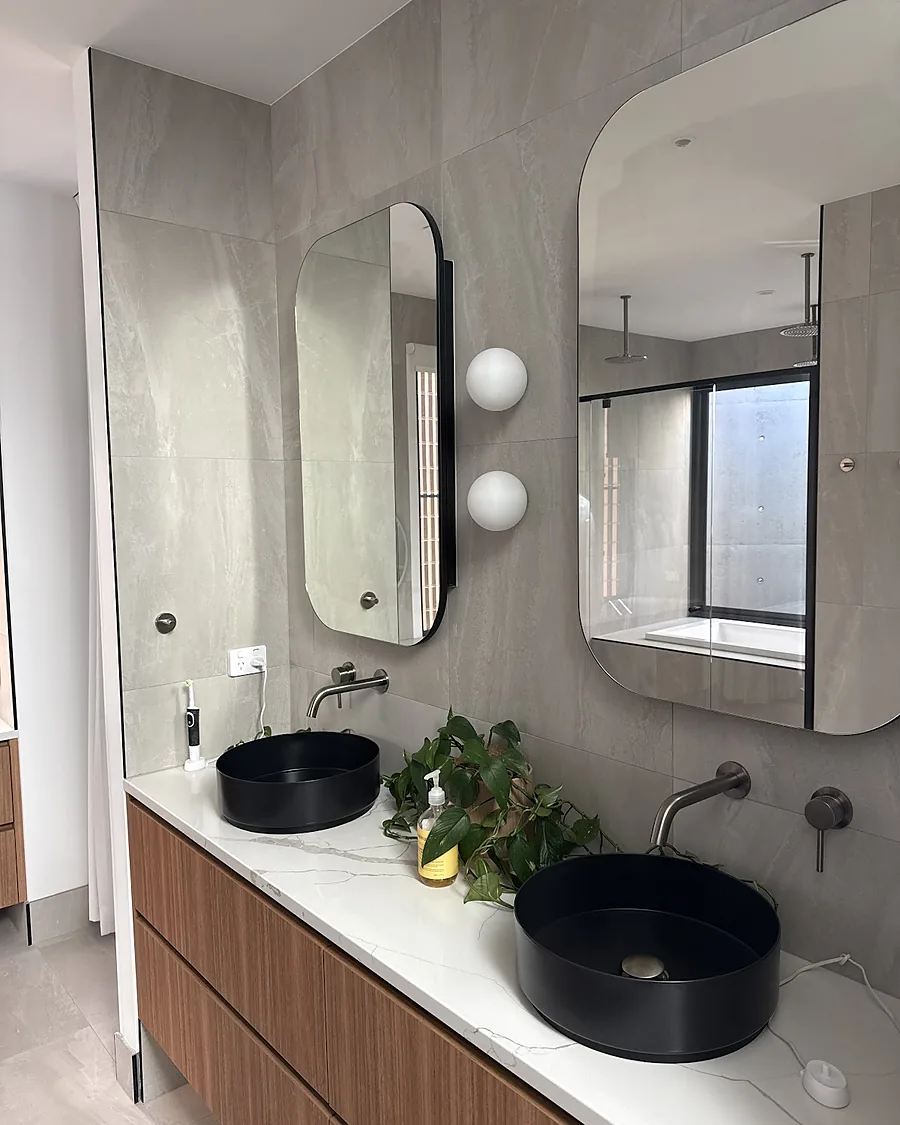 A finished bathroom with a timber vanity, twin black basins, wall mixers and shaped mirrors