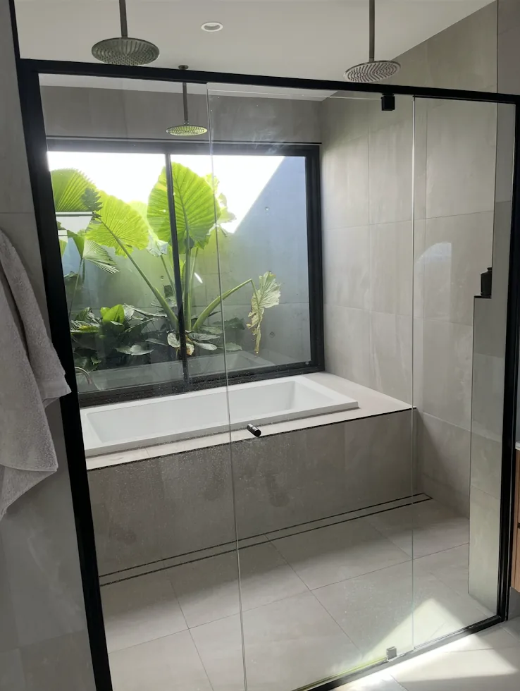 A bath and shower behind a black-framed glass screen with a garden window