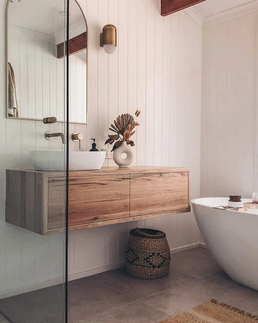 A floating timber vanity with a vessel basin, brass tap, arched mirror and freestanding bath