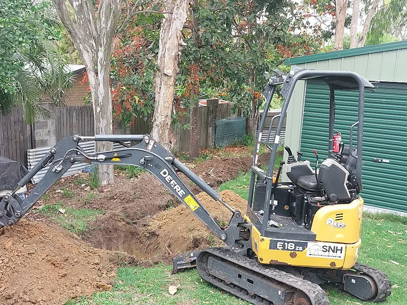 A mini excavator digging a stormwater trench in a backyard beside a green shed
